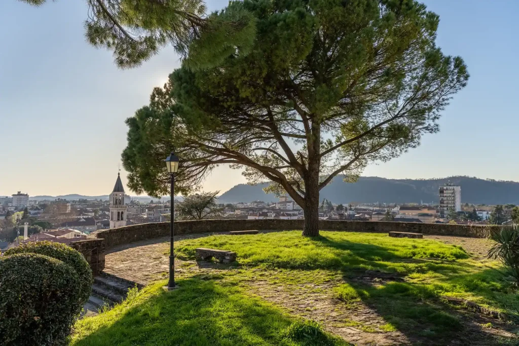 Nell'immagine uno scorcio di Gorizia visto dall'alto. In primo piano c'è uno spiazzo con al centro un albero e delle panchine dal quale vedere il panorama della città